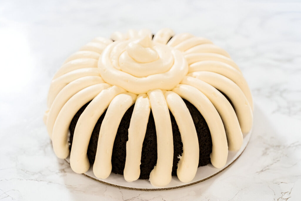 A nothing bundt cake with cream cheese frosting piping on a white cake stand.