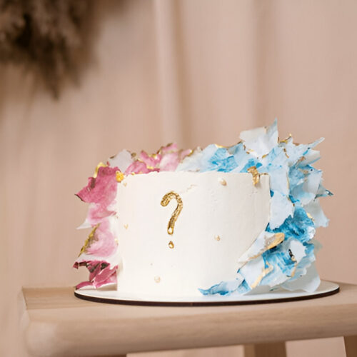 A round white cake with white frosting swirls, topped with question mark written in gold.