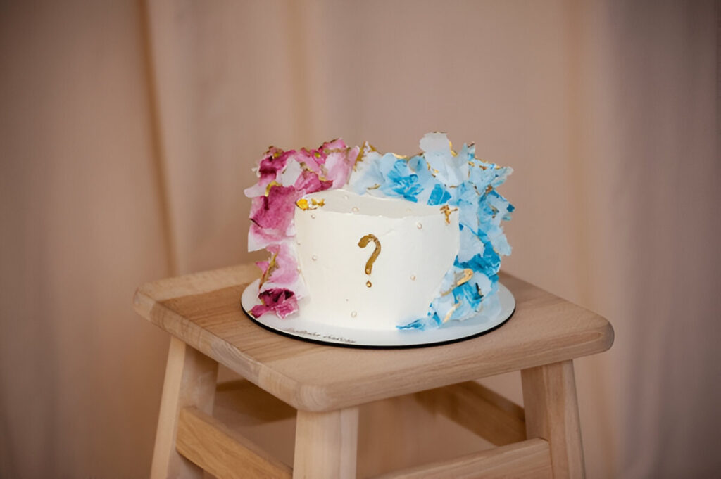 A white frosted gender reveal cake with smooth icing, topped with small pink and blue decorations.